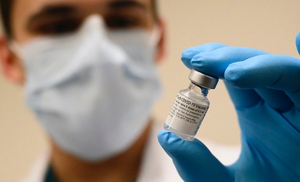 A medical professional wearing a mask holds up a glass vial containing a COVID-19 vaccine.